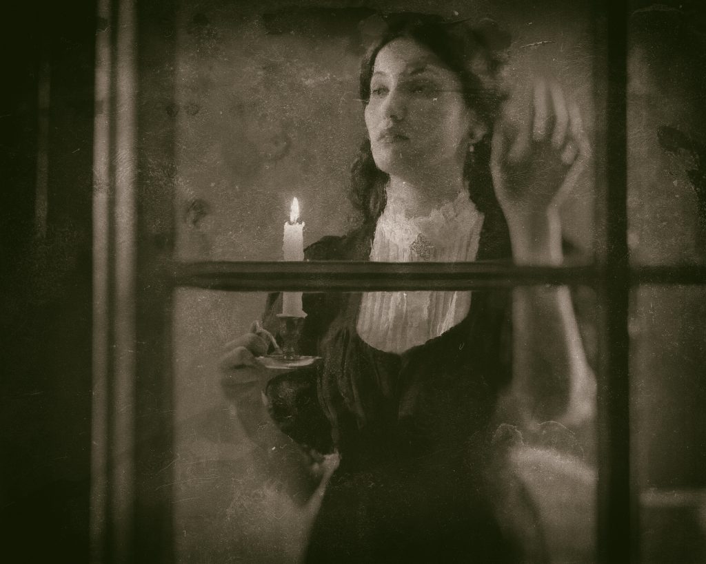 Foto i gråskala av en yngre kvinna i gammaldags kläder som står innanför ett fönster med ett ljus i handen.